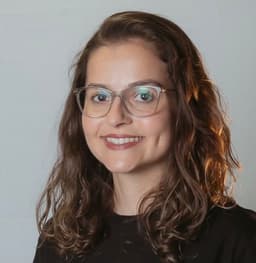 Bruna Nunes -Mulher branca, cabelo castanho claro, ondulado, usando óculos de grau e camiseta preta. Ela está sorrindo.